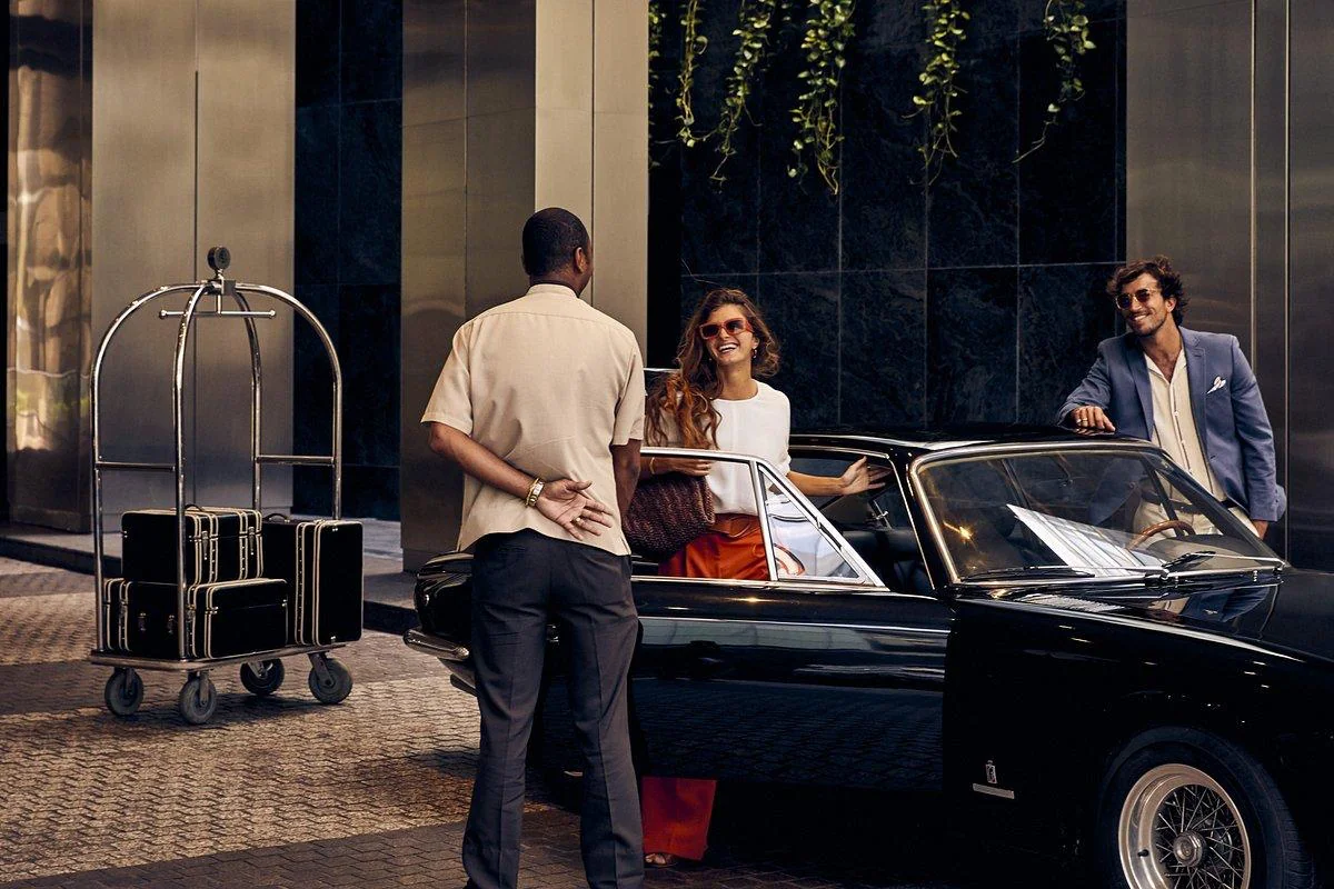 Hotel attendant welcoming guests beside a black convertible and luggage trolley