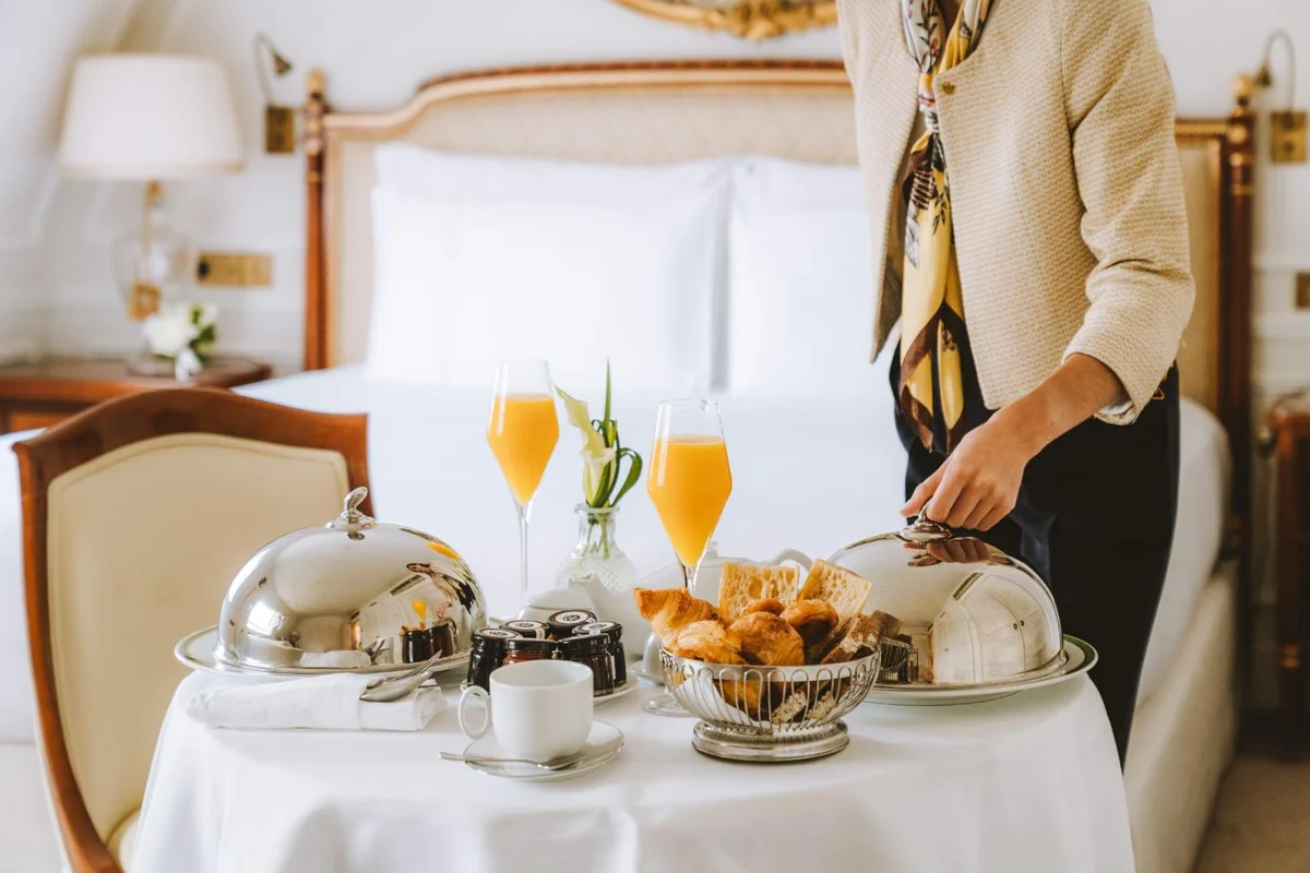Staff member arranging breakfast with pastries and silver serving covers in a hotel suite