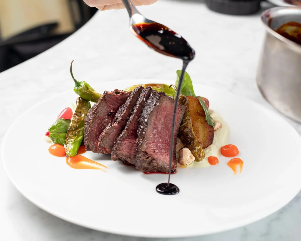 Chef pouring sauce over sliced steak and vegetables on a white plate