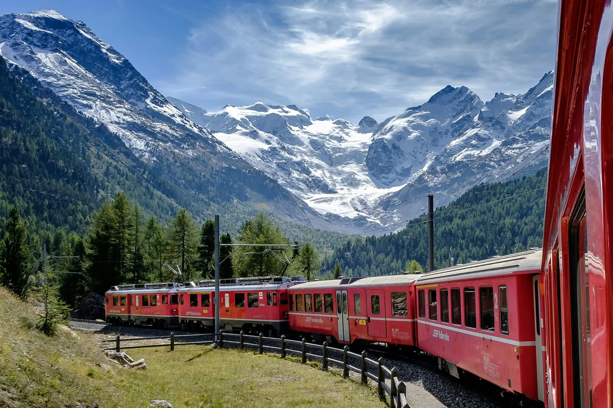 Red passenger train winding through a forest beneath snow-covered mountain peaks